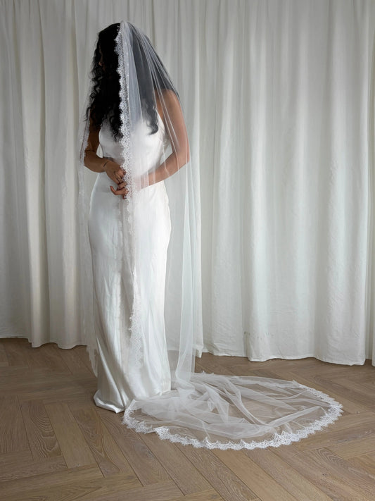 Person wearing a white wedding dress with a long veil in front of white curtains.