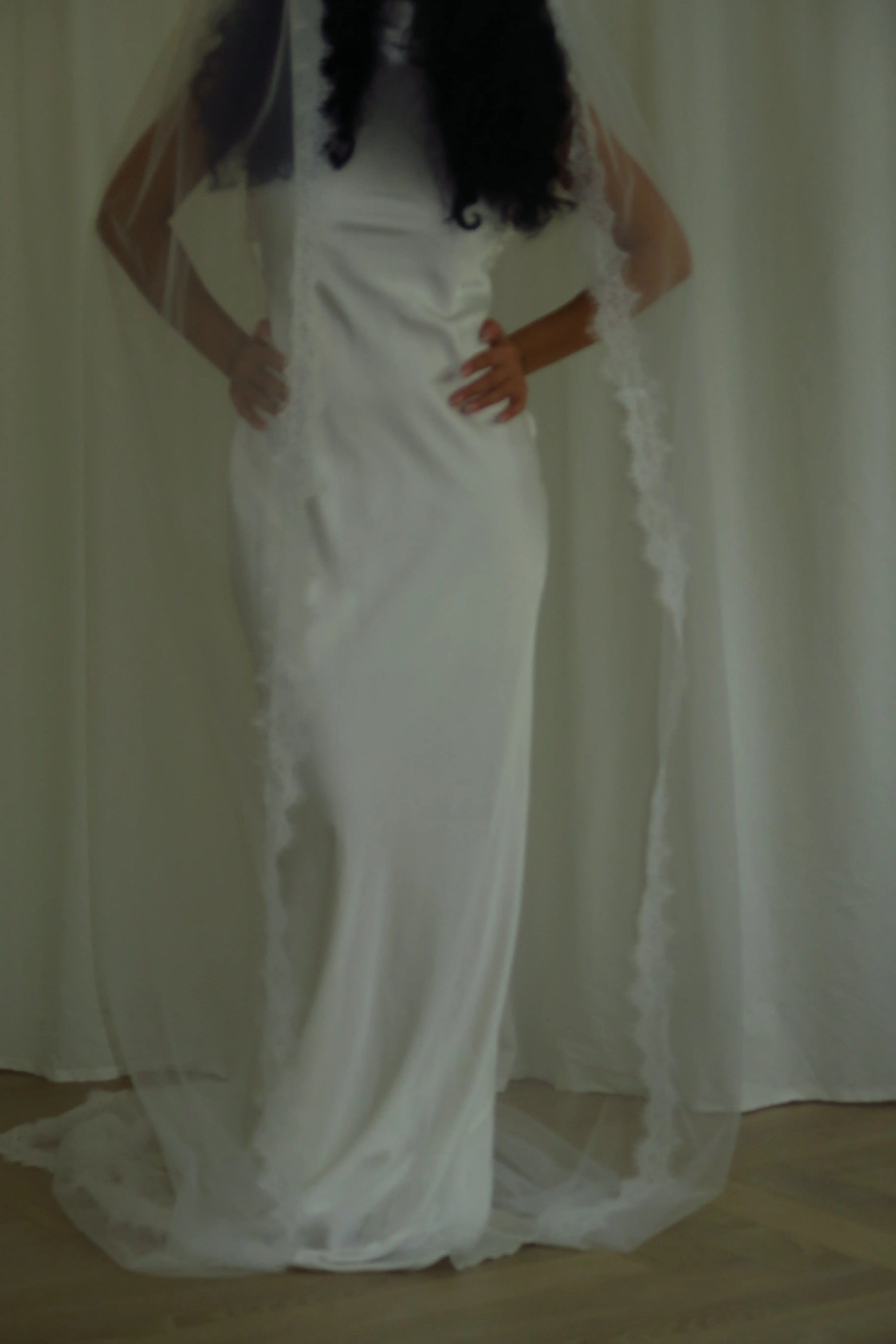 Person wearing a white wedding dress with a long veil in front of white curtains.