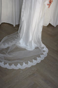 Load image into Gallery viewer, A white bridal veil with a delicate floral lace border draped over a light wood herringbone floor, with a white dress and draped fabric in the background.
