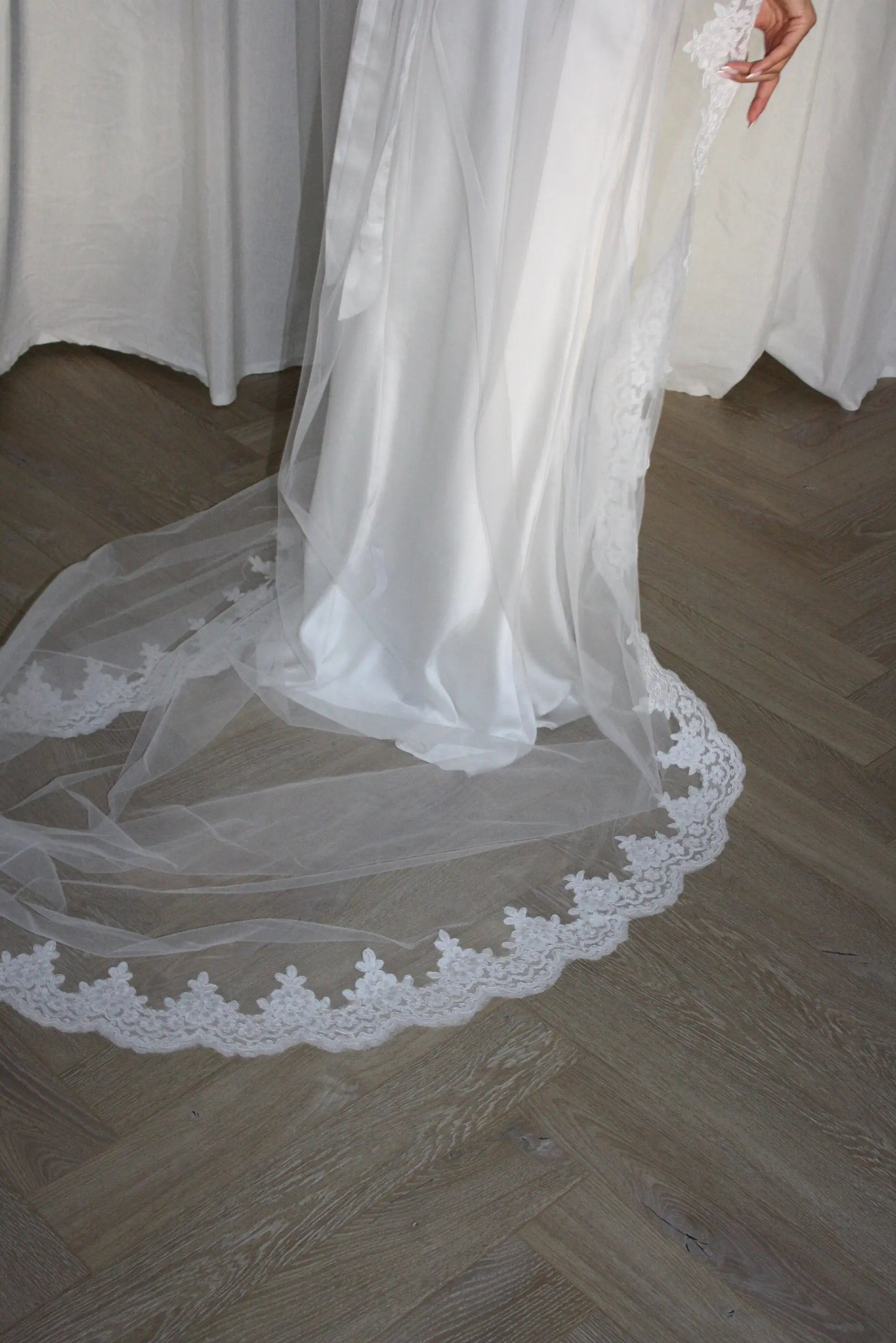A white bridal veil with a delicate floral lace border draped over a light wood herringbone floor, with a white dress and draped fabric in the background.