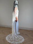 Load image into Gallery viewer, A woman stands in profile wearing a long white lace bridal veil and a wedding dress with a long train, photographed indoors against a plain wall on a wood floor.
