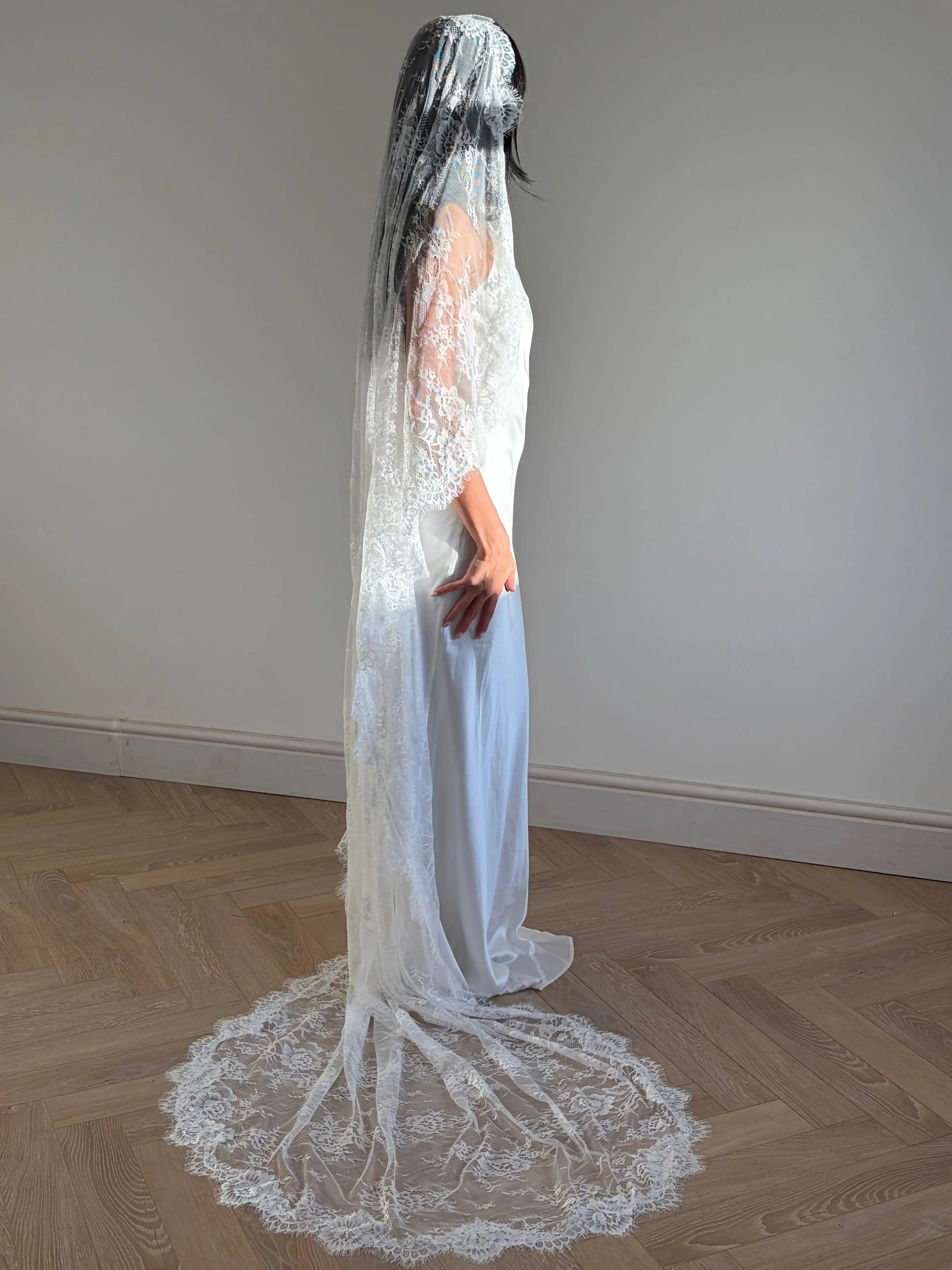 A woman stands in profile wearing a long white lace bridal veil and a wedding dress with a long train, photographed indoors against a plain wall on a wood floor.