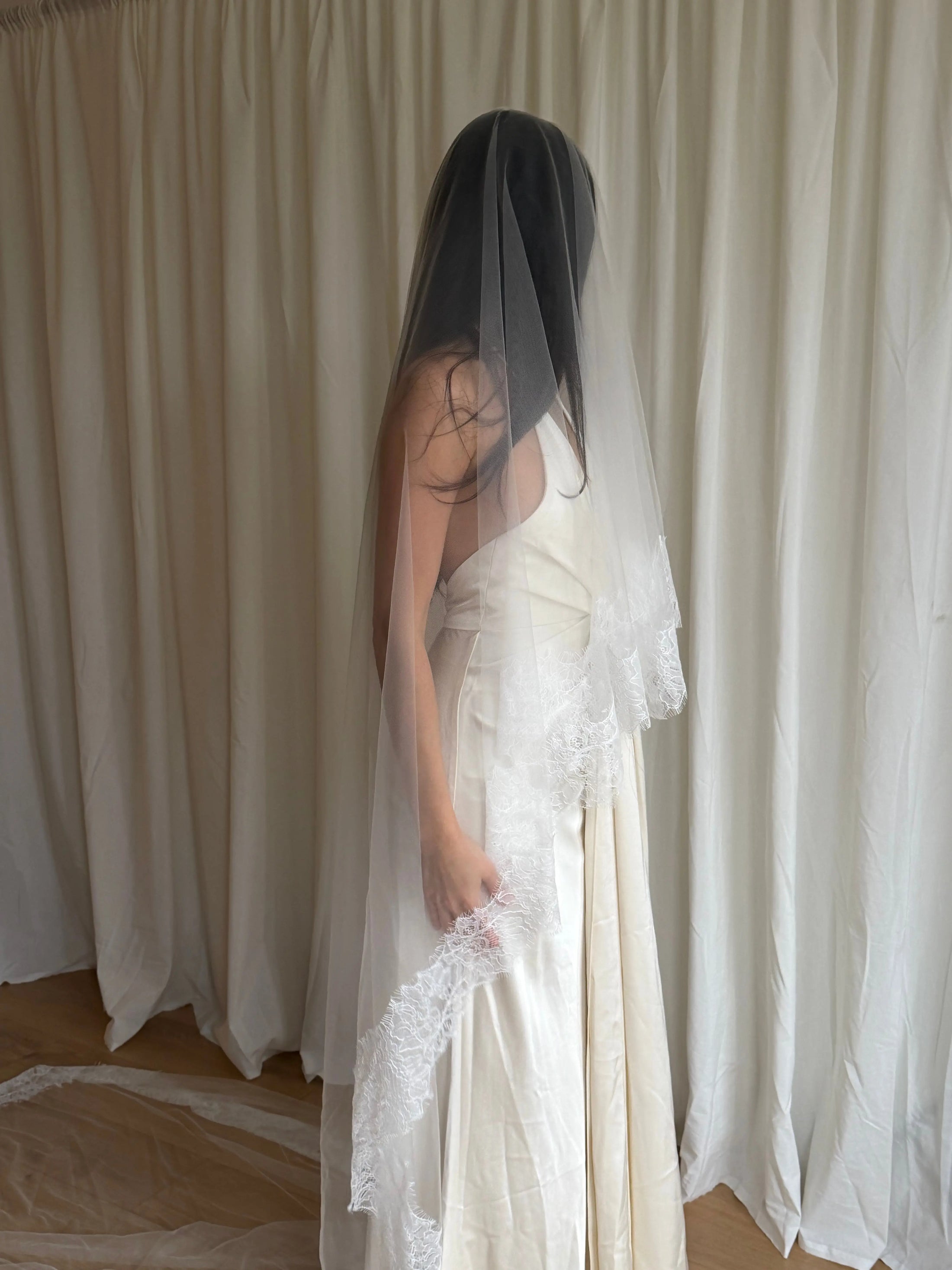 Bride wearing a white wedding dress with a lace veil against a white curtain background.