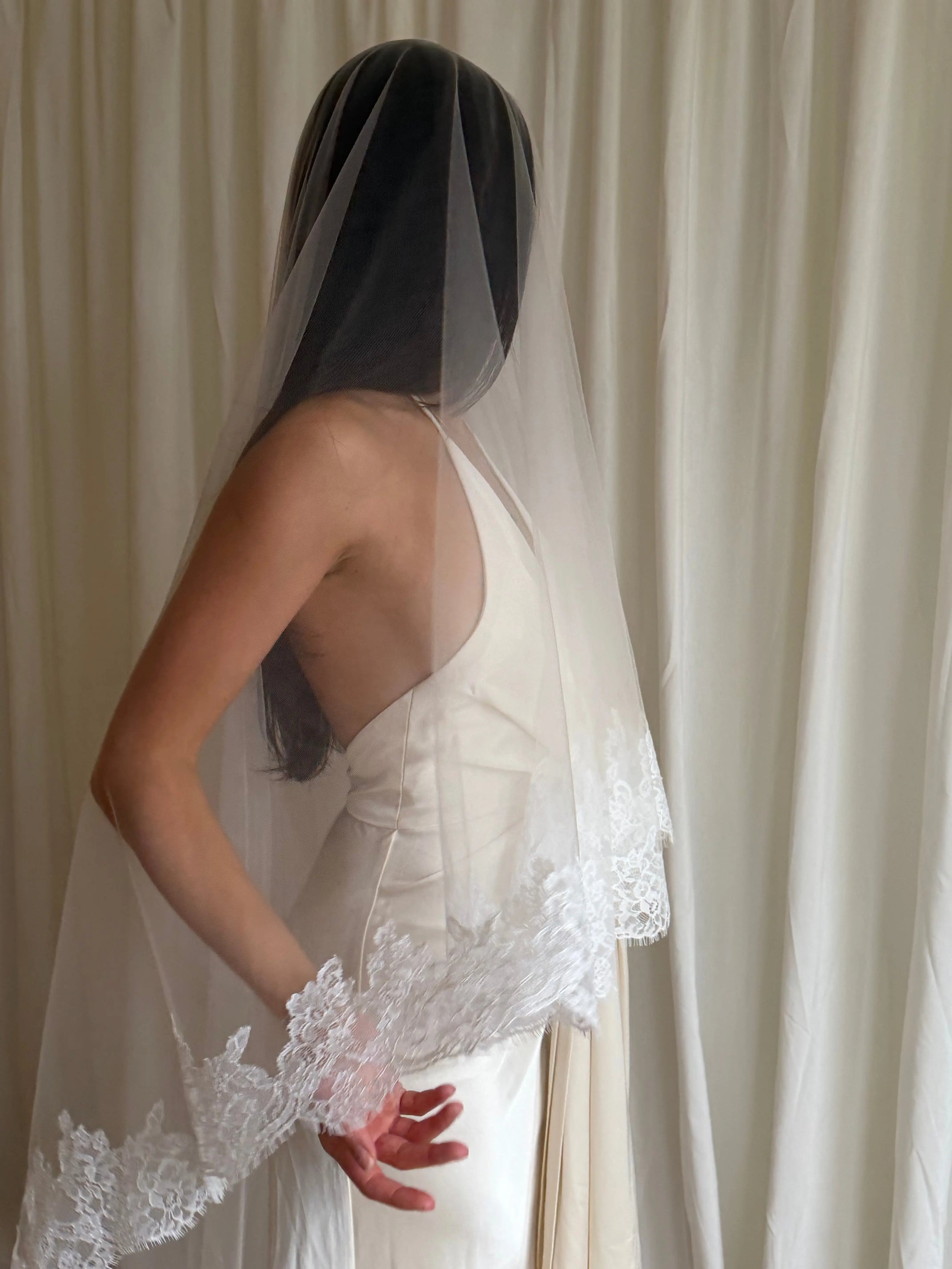 person in a wedding dress with a long lace trim veil against a curtain backdrop.