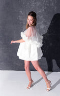 Load image into Gallery viewer, A woman modeling a short white dress with a 3d flower tulle bridal cape standing against a dark textured studio wall.
