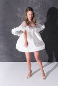 Load image into Gallery viewer, A woman modeling a short white dress with a 3d flower tulle bridal cape standing against a dark textured studio wall.
