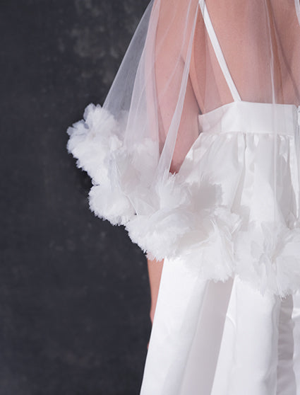 A woman modeling a short white dress with a 3d flower tulle bridal cape standing against a dark textured studio wall.