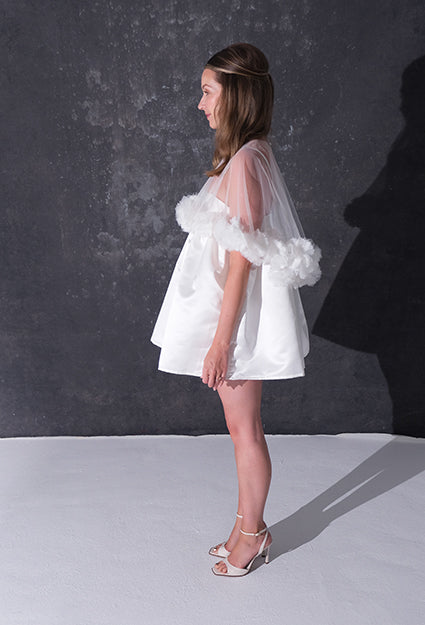 A woman modeling a short white dress with a 3d flower tulle bridal cape standing against a dark textured studio wall.
