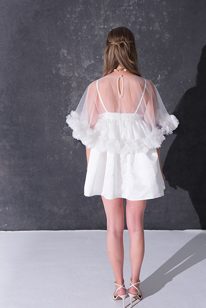 A woman modeling a short white dress with a 3d flower tulle bridal cape standing against a dark textured studio wall.