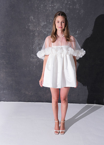 A woman modeling a short white dress with a 3d flower tulle bridal cape standing against a dark textured studio wall.