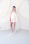 Load image into Gallery viewer, A woman wearing a sheer white bridal veil with delicate bow details and a short white slip dress, standing in front of draped white fabric.
