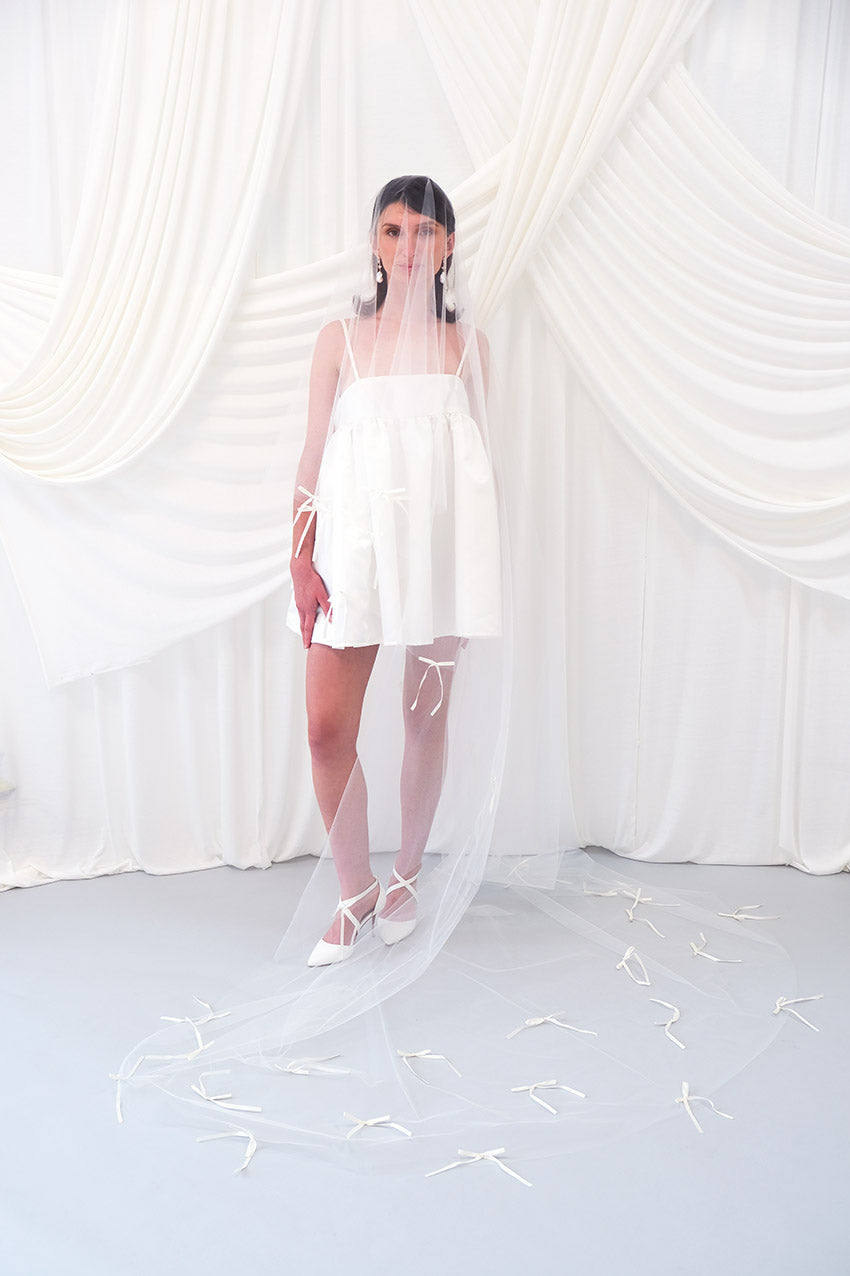 A woman wearing a sheer white bridal veil with delicate bow details and a short white slip dress, standing in front of draped white fabric.