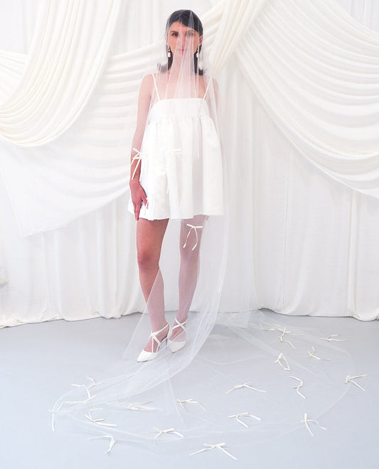 A woman wearing a sheer white bridal veil with delicate bow details and a short white slip dress, standing in front of draped white fabric.