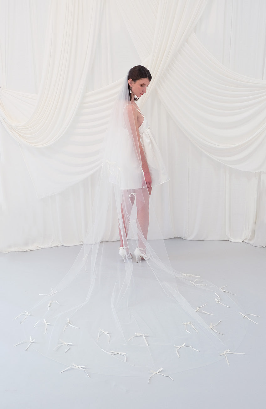 A woman wearing a sheer white bridal veil with delicate bow details and a short white slip dress, standing in front of draped white fabric.