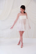 Load image into Gallery viewer, A woman wearing a sheer white bridal veil with delicate bow details and a short white slip dress, standing in front of draped white fabric.

