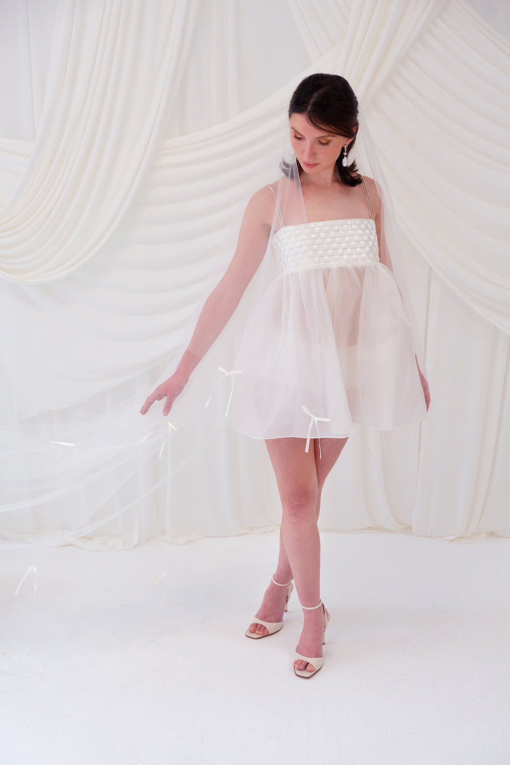 A woman wearing a sheer white bridal veil with delicate bow details and a short white slip dress, standing in front of draped white fabric.