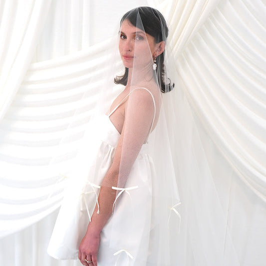 A woman wearing a sheer white bridal veil with delicate bow details and a short white slip dress, standing in front of draped white fabric.