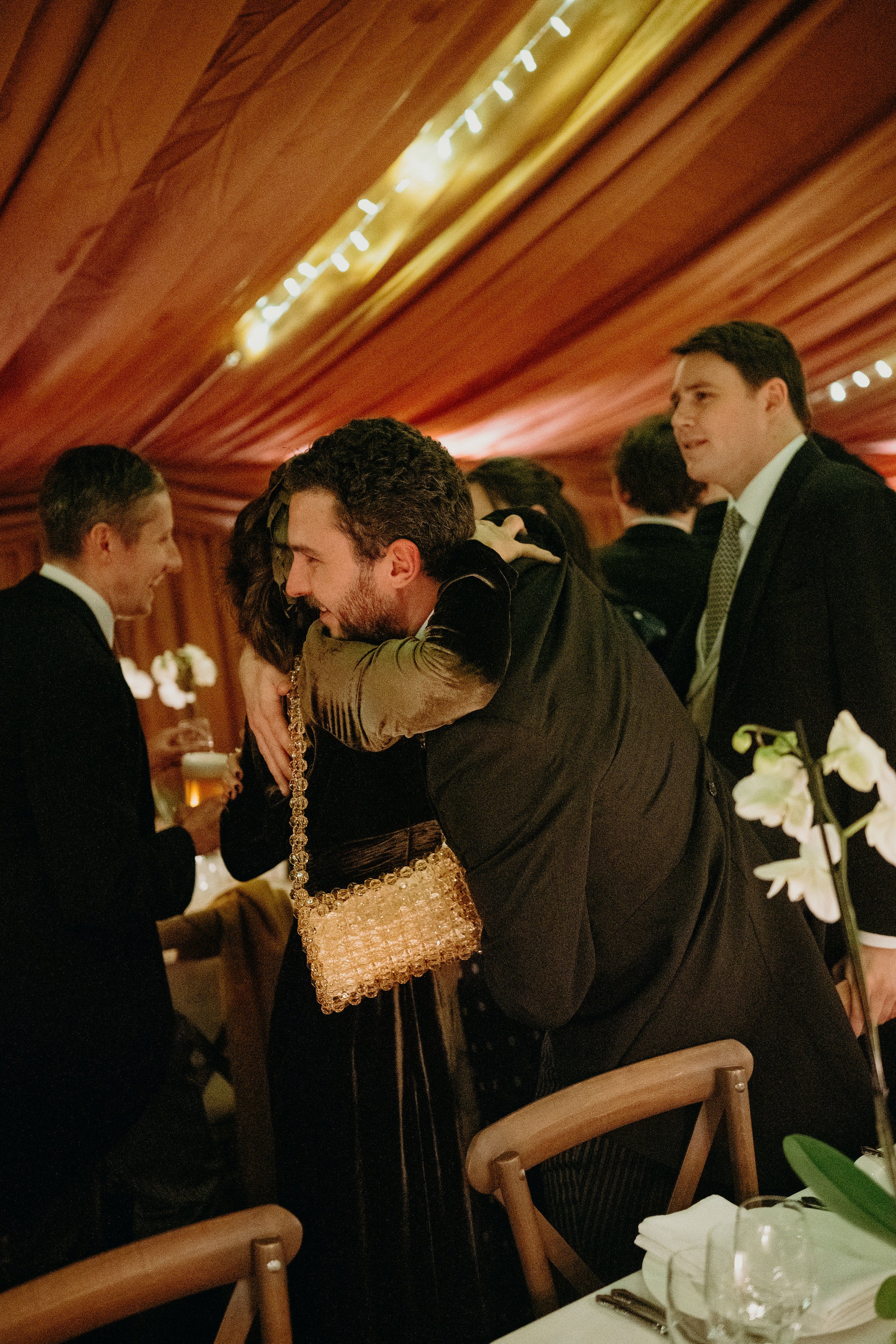 Two people embracing at a formal event with wooden ceiling and string lights wearing bridal bag