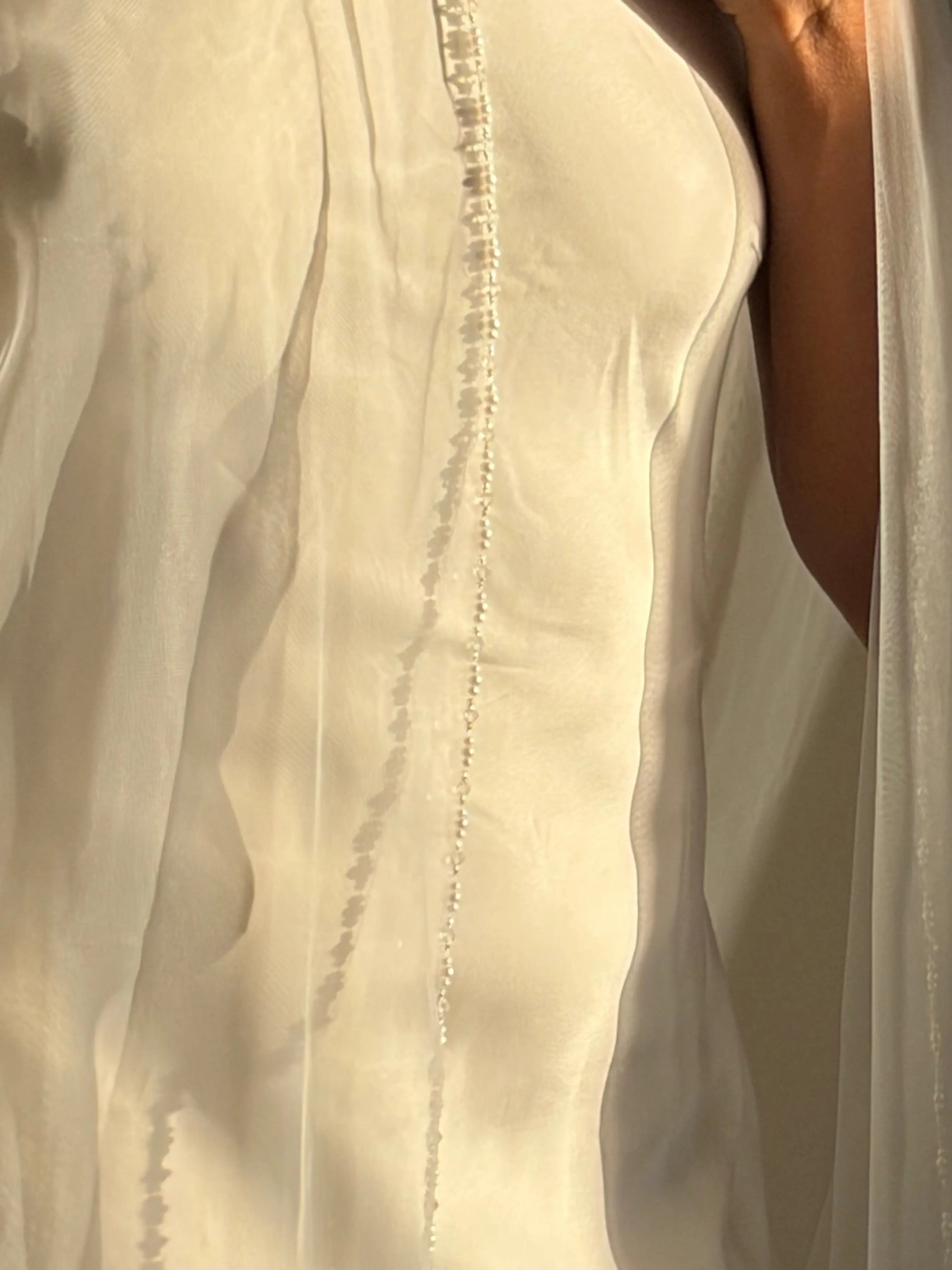Close-up of a white bridal veil or dress fabric with delicate bead trim and a warm-toned model's shoulder visible on the right.