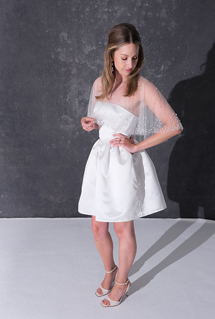 A woman modeling a short white dress with a pearl tulle bridal cape standing against a dark textured studio wall.