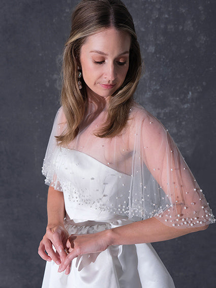 A woman modeling a short white dress with a pearl tulle bridal cape standing against a dark textured studio wall.