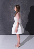 Load image into Gallery viewer, A woman modeling a short white dress with a pearl tulle bridal cape standing against a dark textured studio wall.
