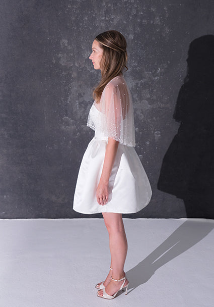 A woman modeling a short white dress with a pearl tulle bridal cape standing against a dark textured studio wall.