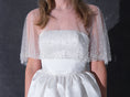 Load image into Gallery viewer, A woman modeling a short white dress with a pearl tulle bridal cape standing against a dark textured studio wall.
