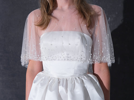 A woman modeling a short white dress with a pearl tulle bridal cape standing against a dark textured studio wall.