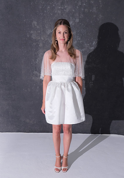 A woman modeling a short white dress with a pearl tulle bridal cape standing against a dark textured studio wall.