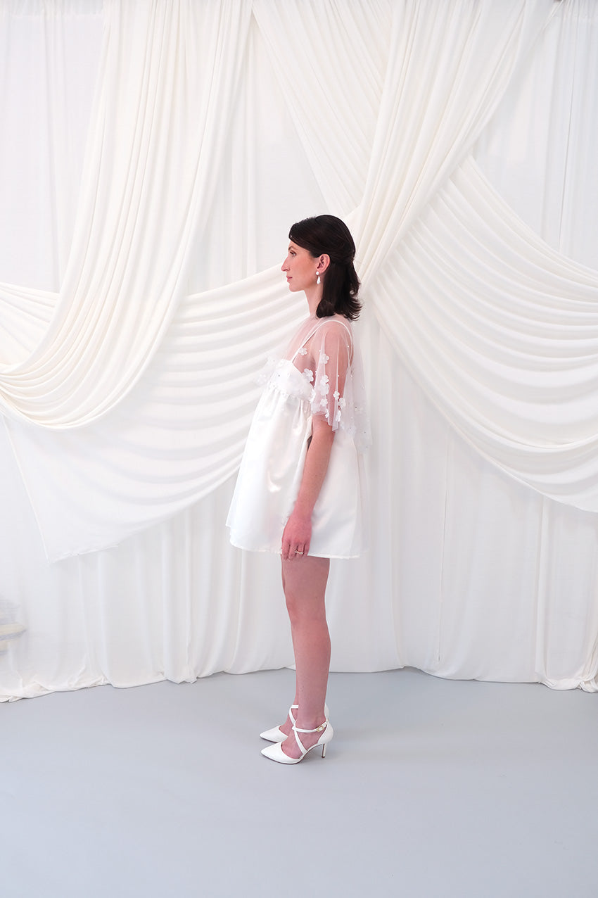 A young girl modeling a short white slip dress with a tulle petal bridal cape and white pointed-toe heels, standing against a draped white fabric backdrop.