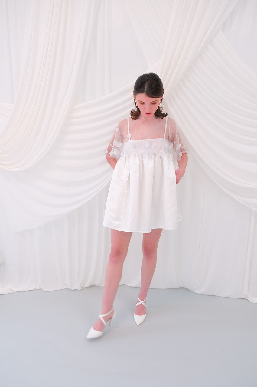 A young girl modeling a short white slip dress with a tulle petal bridal cape and white pointed-toe heels, standing against a draped white fabric backdrop.