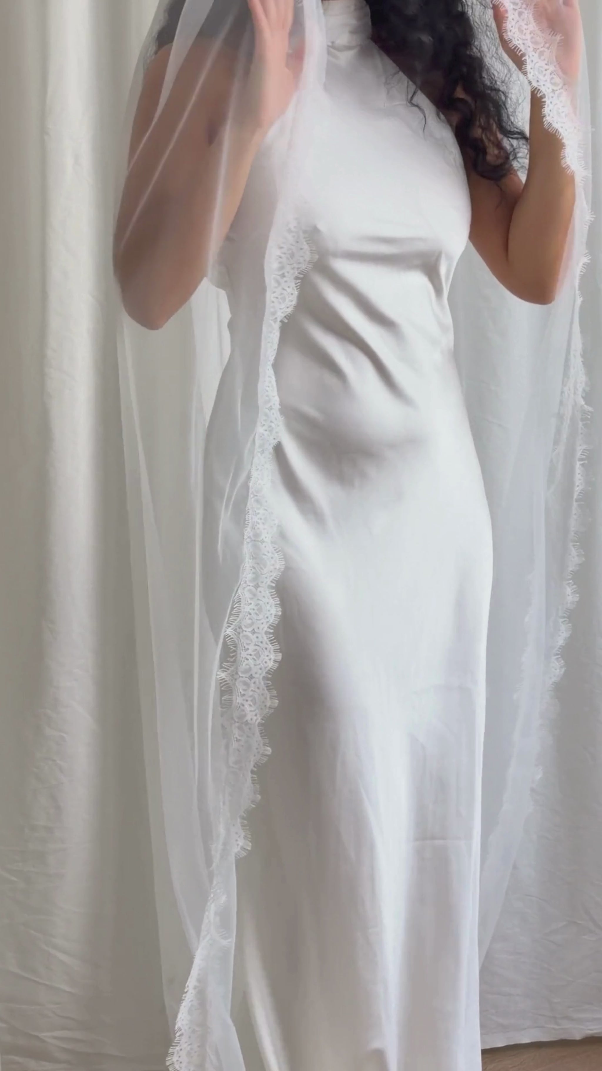 Person wearing a white wedding dress with a long veil in front of white curtains.