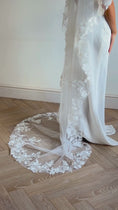 Load and play video in Gallery viewer, A woman wearing a long white bridal veil with floral lace appliqué and a matching sleeveless wedding dress, standing in a room with a light beige wall and wood herringbone floor.

