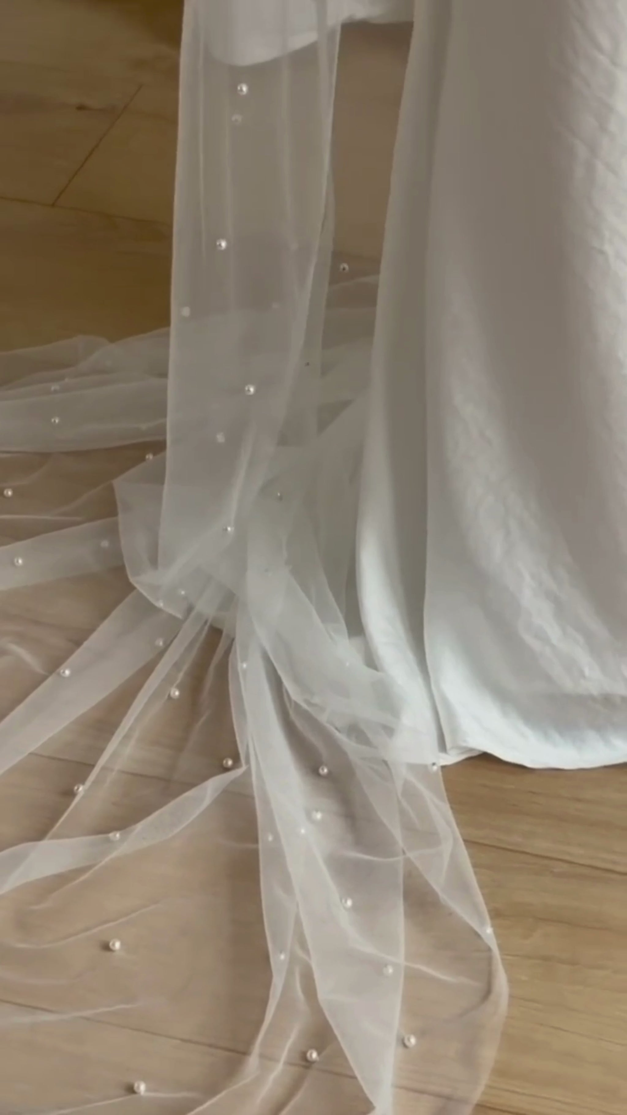 Person wearing a white wedding dress with a long pearl veil against a plain background