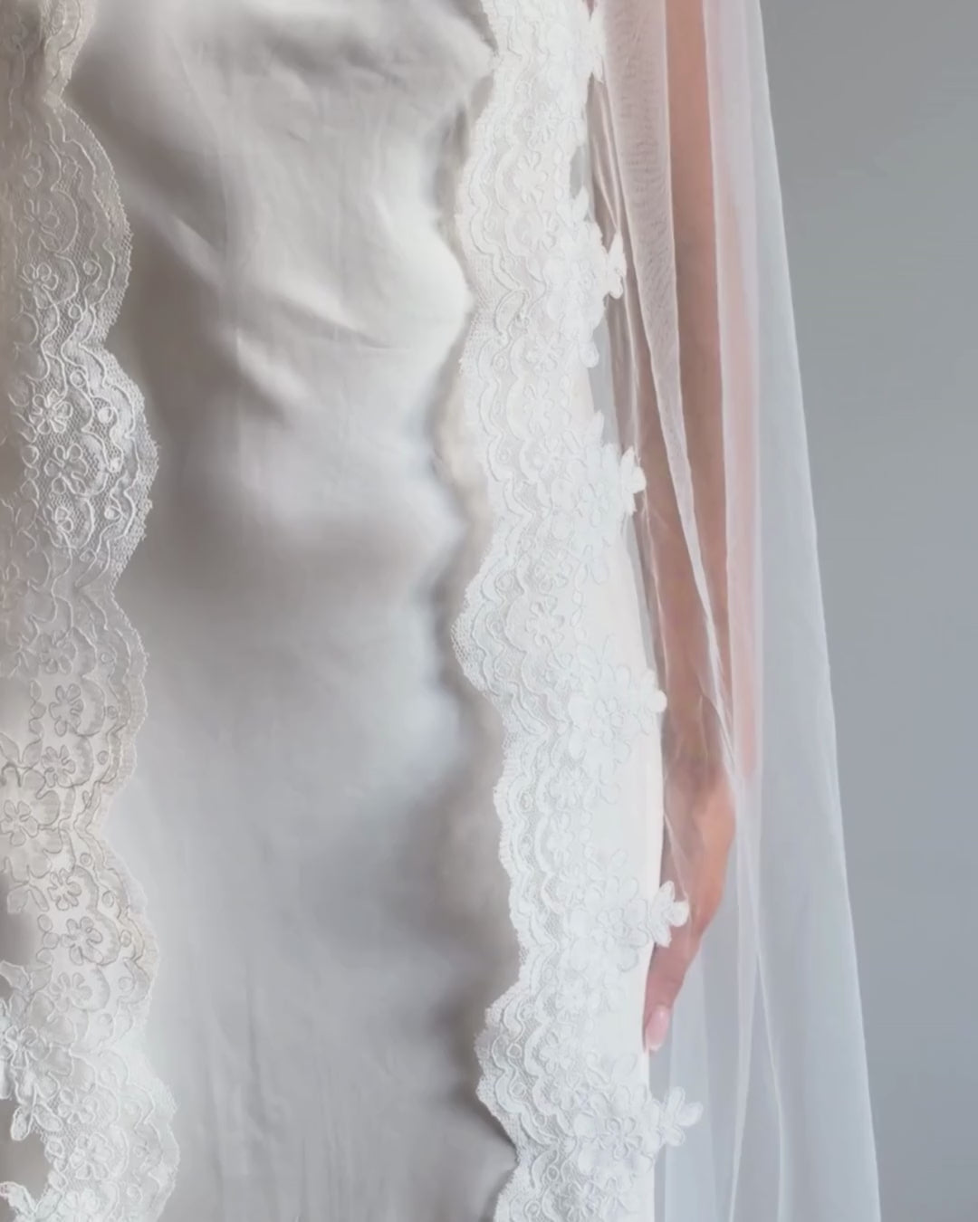 A woman in a white wedding dress and long sheer veil with lace trim stands in profile in a room with light wood herringbone flooring and a plain off-white wall.