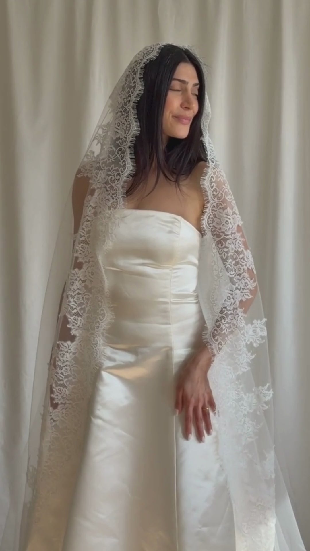 Bride wearing a white wedding dress with a lace veil against a white curtain background.