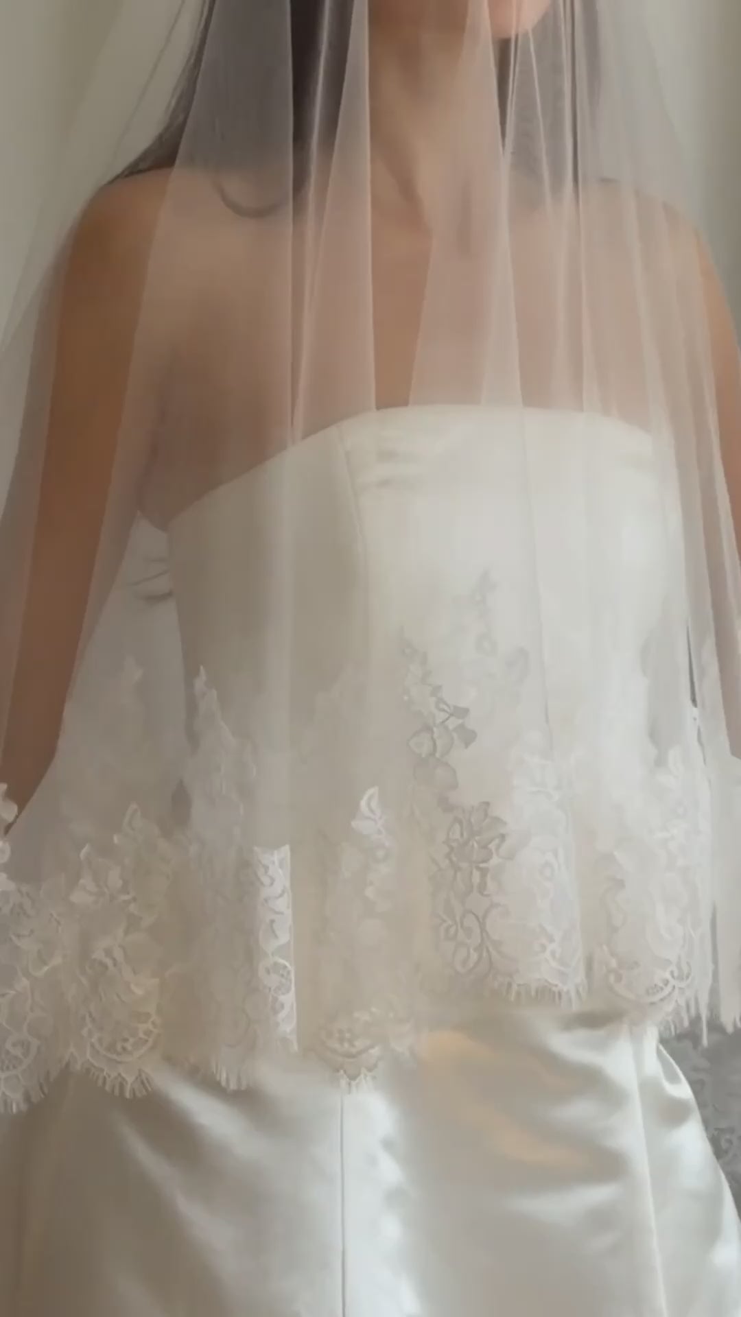 Bride wearing a white wedding dress with a lace veil against a white curtain background.