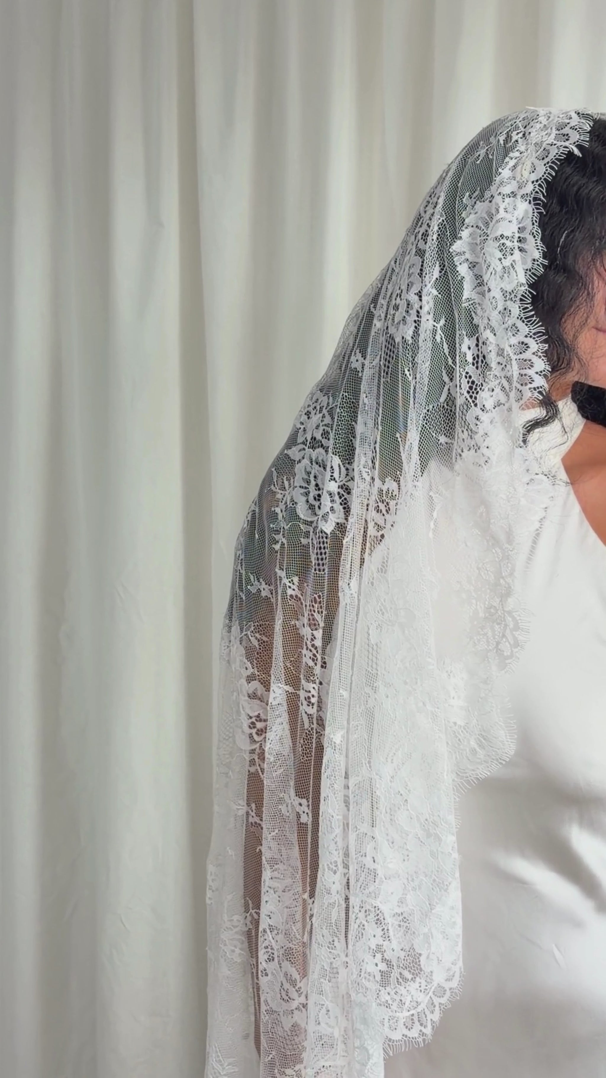 Person wearing a white wedding dress with a long lace veil against a plain background