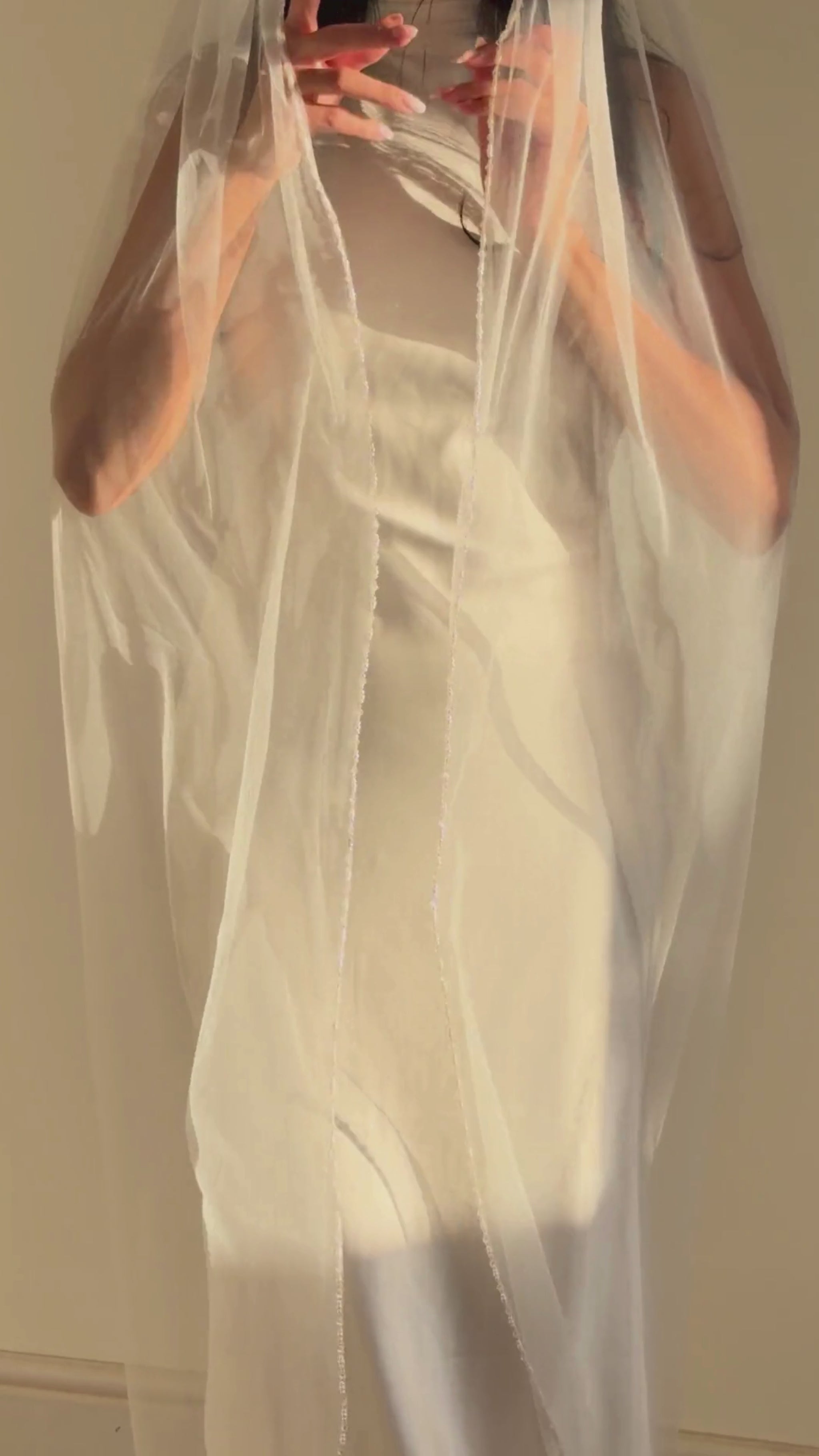 Person wearing a white wedding dress with a long pearl veil against a plain background