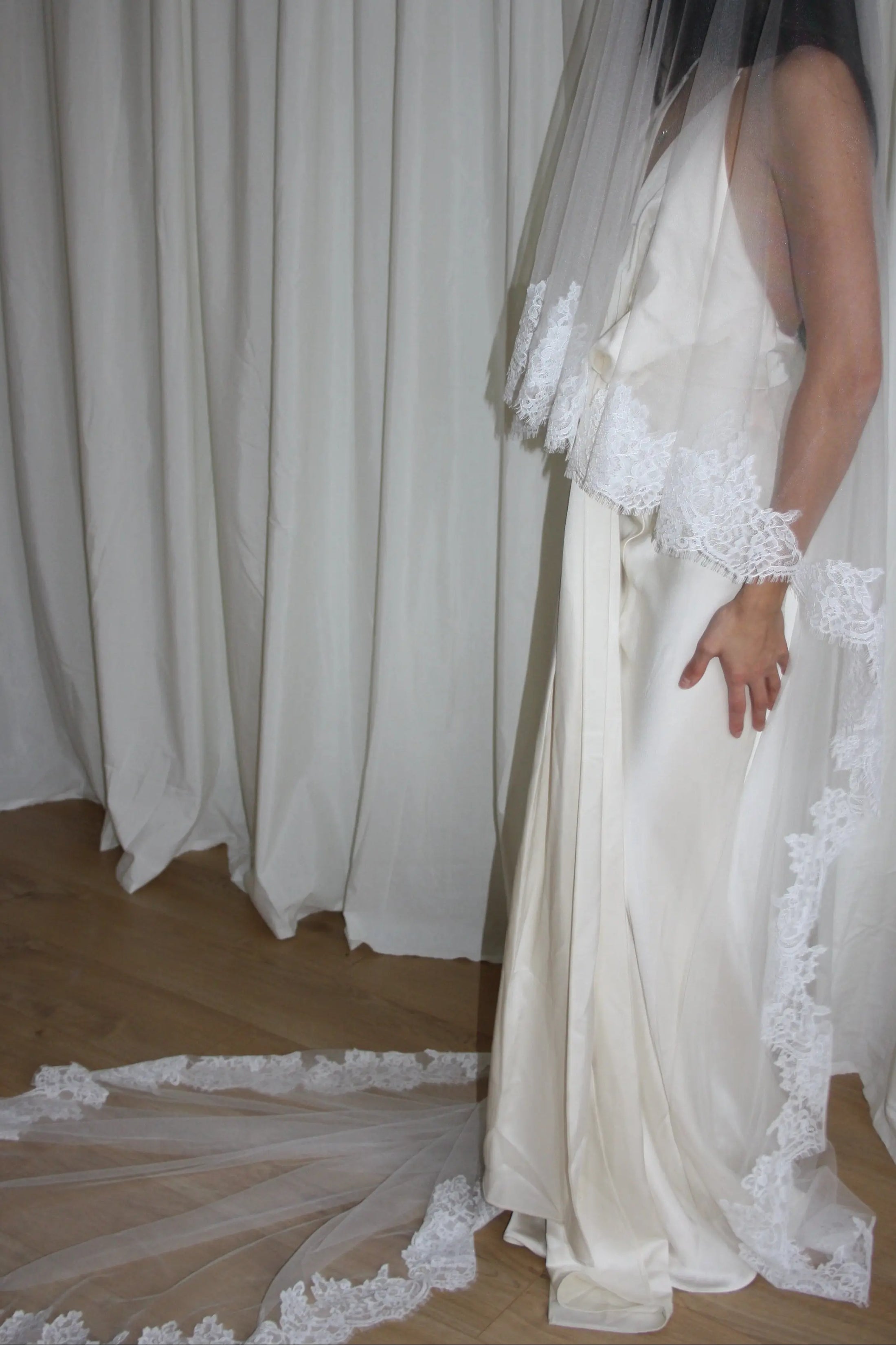 person in a wedding dress with a long lace trim veil against a curtain backdrop.