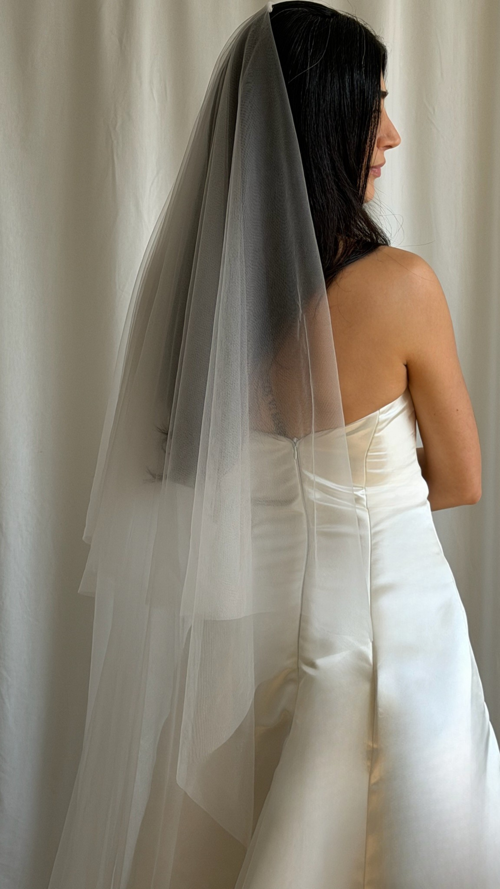 Bride wearing a white wedding dress with a plain veil against a white curtain background.