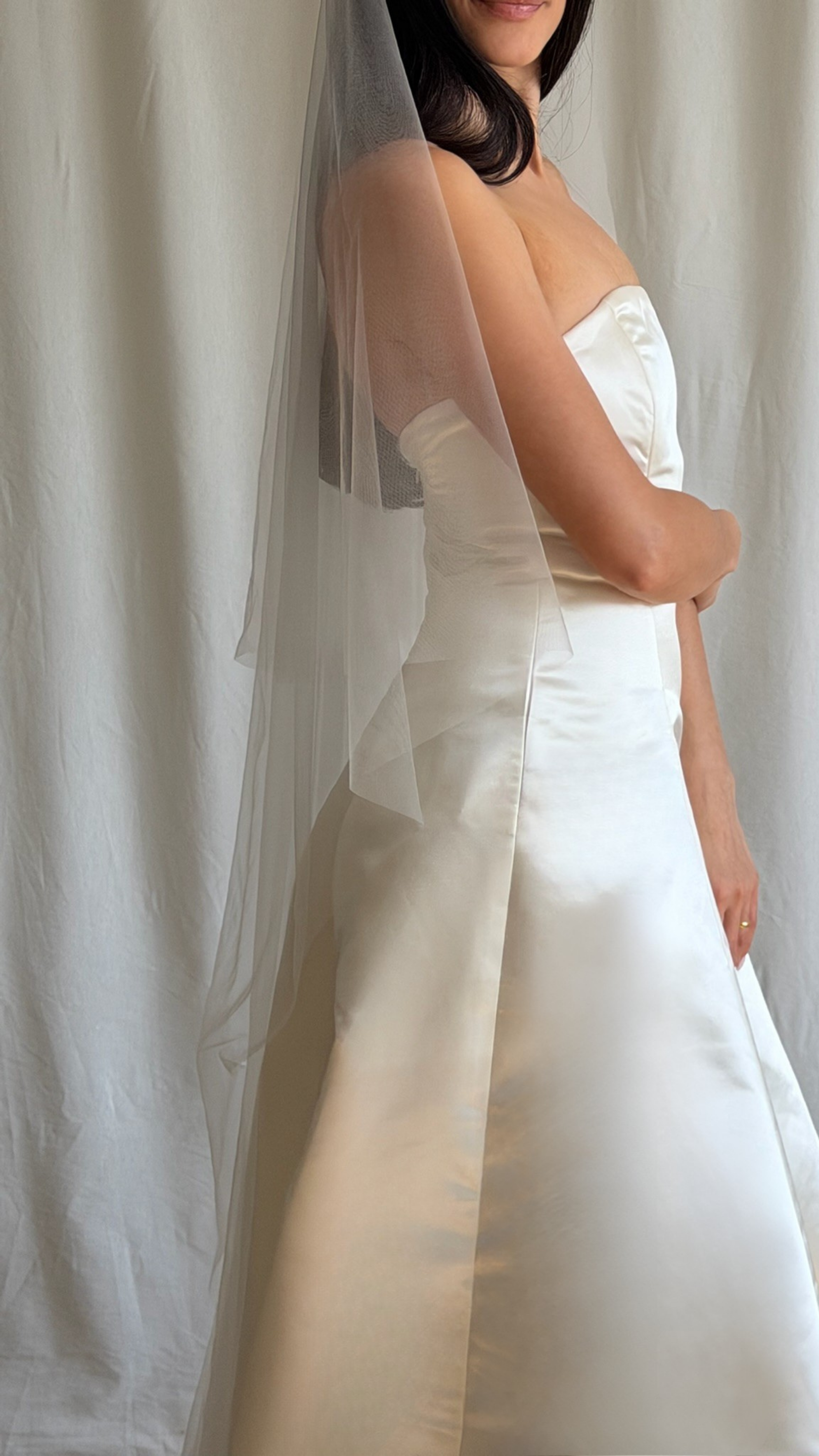 Bride wearing a white wedding dress with a plain veil against a white curtain background.