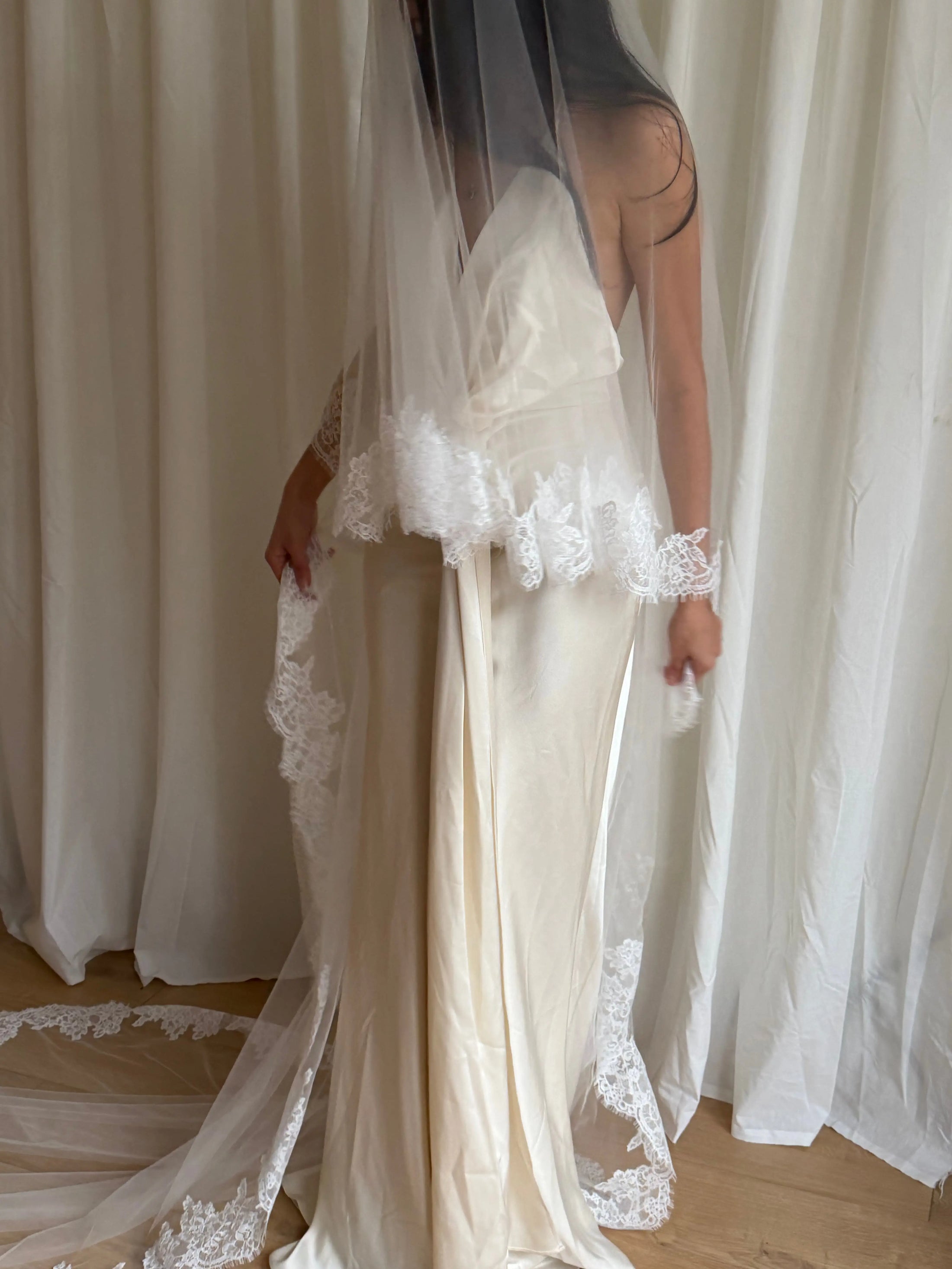 person in a wedding dress with a long lace trim veil against a curtain backdrop.