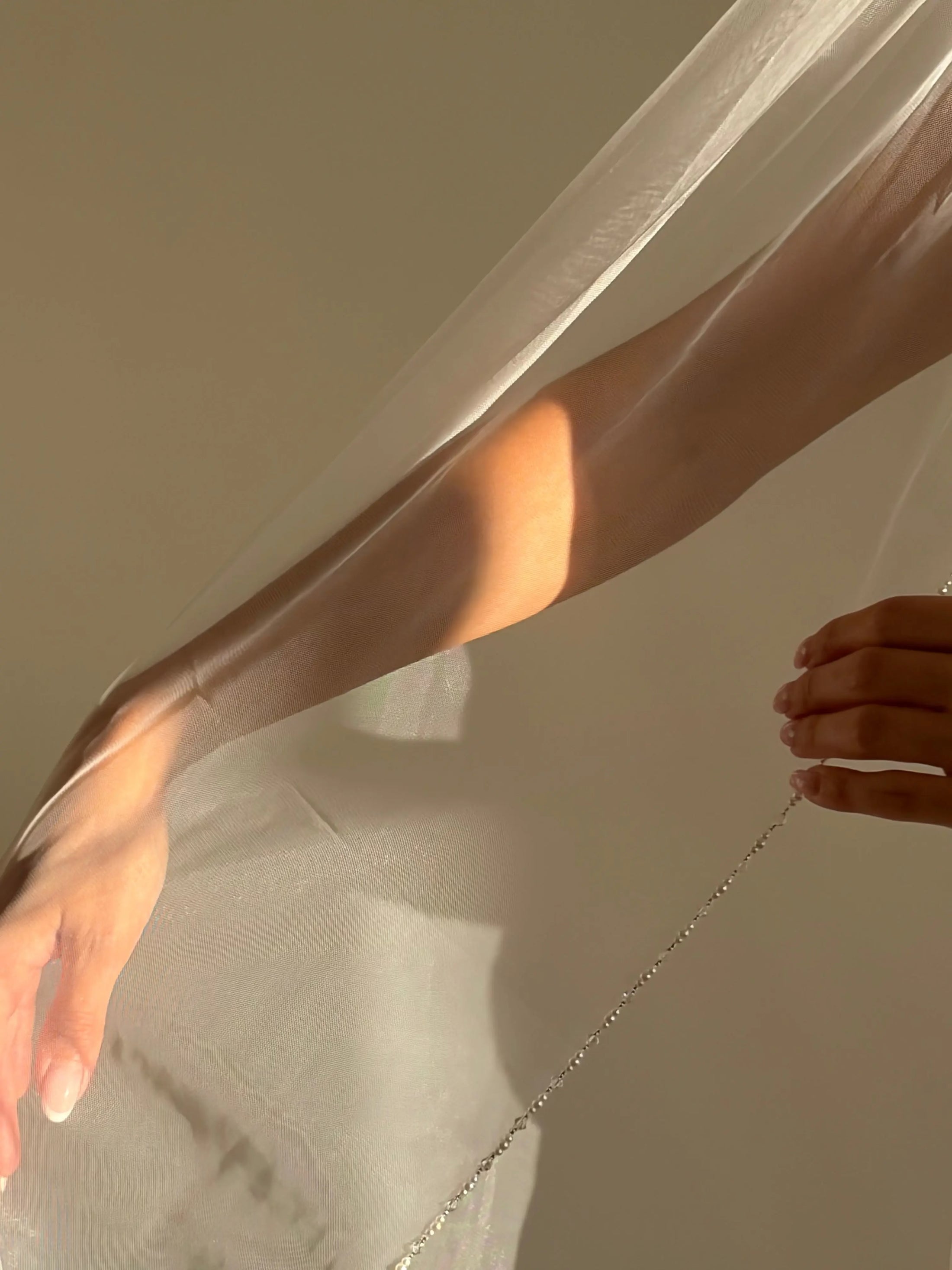 A close-up of a person's arm and hand holding a sheer white veil or tulle fabric, with warm sunlight casting shadows across the skin and fabric against a plain beige background.