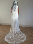Load image into Gallery viewer, A woman viewed from behind wearing a long white lace bridal veil over her head and shoulders, standing on a light wood herringbone floor against a plain off-white wall.
