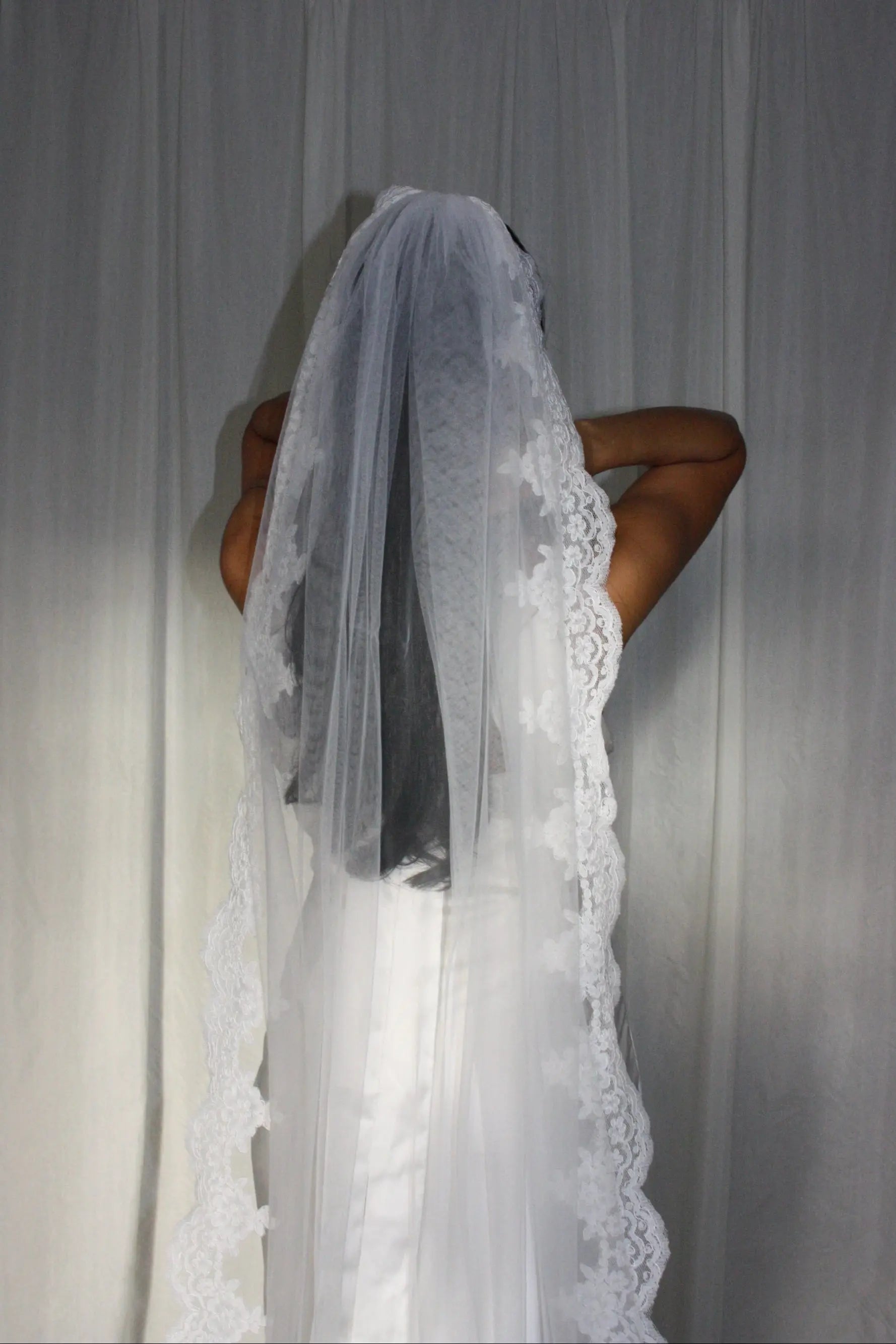 A long white lace bridal veil worn by a model, shown from the back against a draped white fabric backdrop.