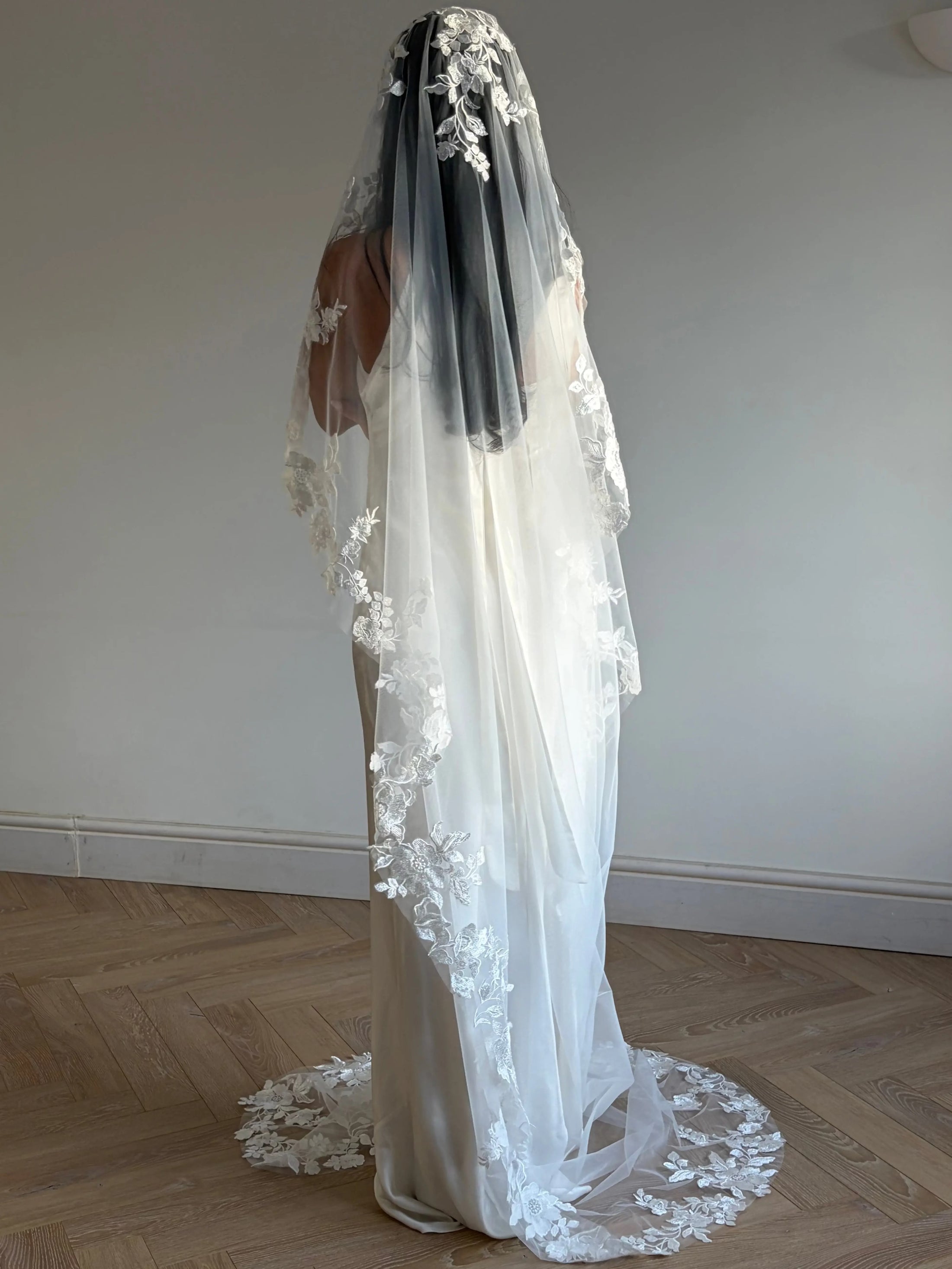 A woman wearing a long white lace bridal veil over her head and shoulders, paired with a white wedding dress, standing on a light wood herringbone floor against a plain off-white wall.