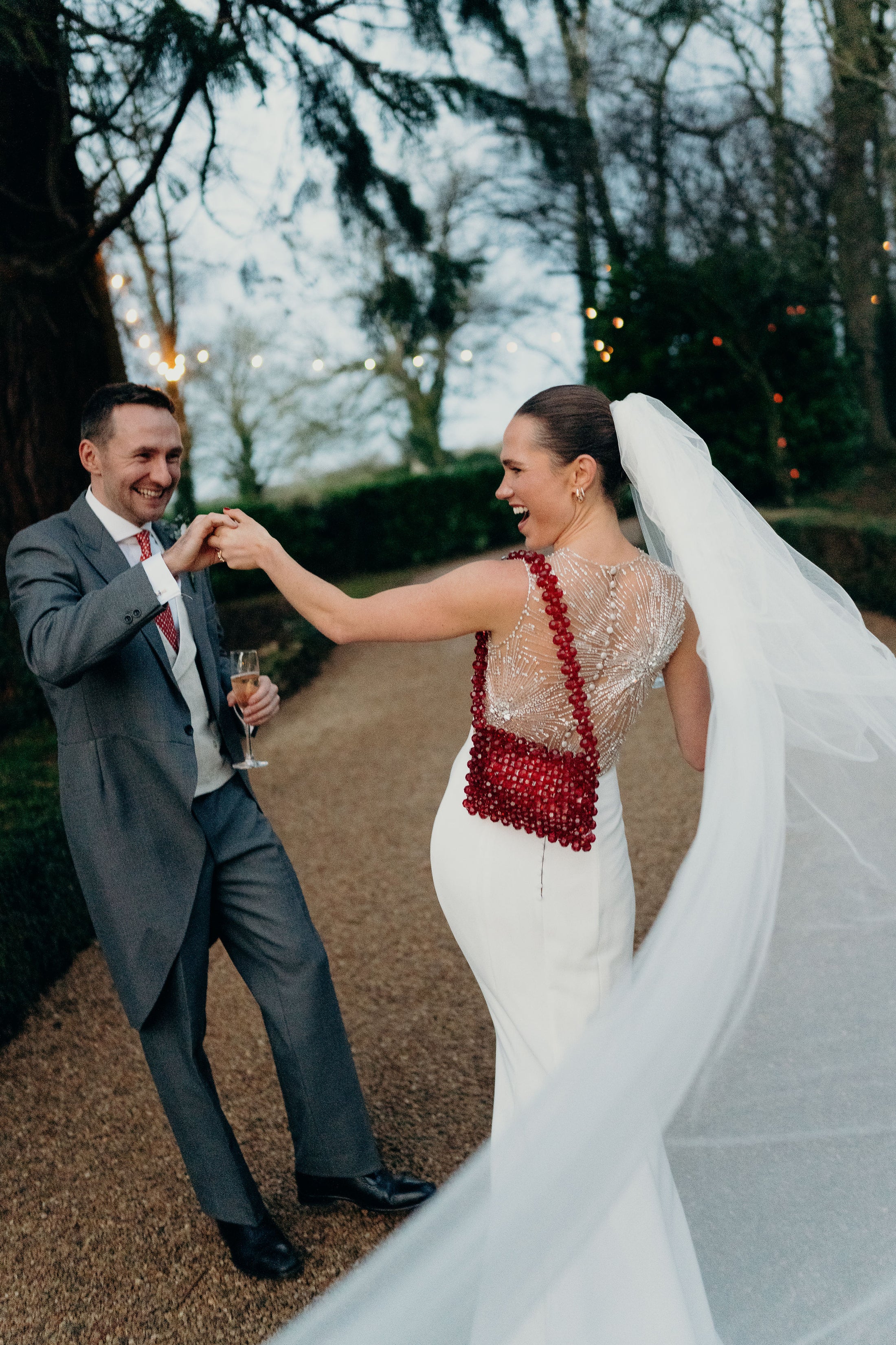 Man and woman in wedding attire standing outdoors with trees and lights in the background wearing bridal bag